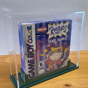 Puede incluir: Una vitrina de plástico transparente con una base verde, que muestra un cartucho de juego de Game Boy Color. El juego es Rugrats: Totally Angelica, con una imagen de dibujos animados de Angelica Pickles en la parte delantera. El juego está clasificado como E para todos.