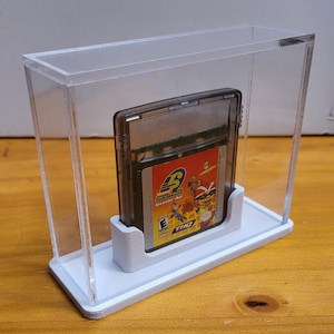 May include: A clear acrylic display case with a white base, showcasing a Game Boy Advance game cartridge. The game cartridge is for "Rocket Power: Beach Bandits" and features a colourful design with the game title and characters.