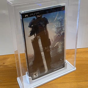 May include: A clear acrylic display case with a white base, showcasing a video game case for the game "Crisis Core: Final Fantasy VII" for the PlayStation Portable (PSP). The game case features a character holding a sword and the game title in gold lettering.