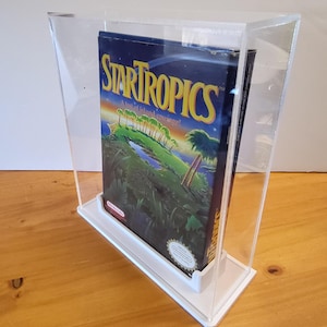 May include: A clear acrylic display case with a white base, showcasing a vintage Nintendo Entertainment System game cartridge box for StarTropics. The box features a colorful illustration of a tropical island with palm trees and a sunset.