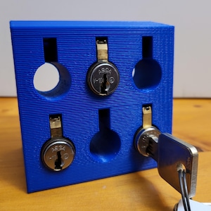 以下が含まれることがあります： 6つのキーシリンダー用のスロットが付いた青いプラスチック製のホルダー。2つのキーシリンダーが見え、どちらも銀色で真鍮のキーが付いています。キーシリンダーには「ASGA V-10」とラベル付けされています。銀色のキーがキーシリンダーの1つに挿入されています。