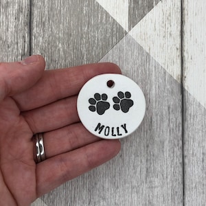 May include: White ceramic dog tag with two black paw prints and the name "MOLLY" written in black.