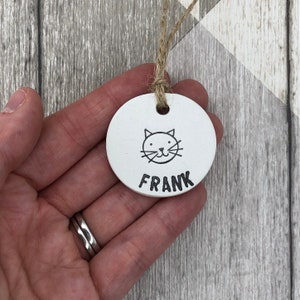 May include: A white, circular ceramic pet tag featuring a cat face illustration and the name "FRANK" in black. It is suspended by a natural twine cord. The background shows a light grey and white wood grain pattern.