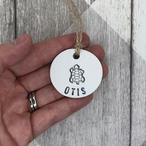 May include: White ceramic ornament featuring a turtle design and the name "OTIS" inscribed below. The ornament is suspended from a string.