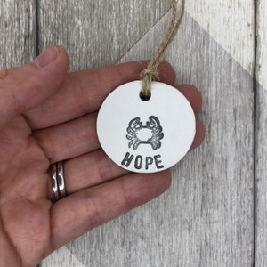 May include: White ceramic ornament featuring a crab design and the word "HOPE" underneath. The ornament is attached to a twine string.