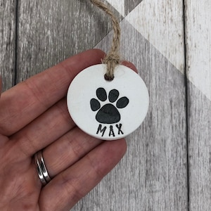 May include: White ceramic ornament with a black paw print and the name "MAX" written underneath. The ornament is attached to a twine string.