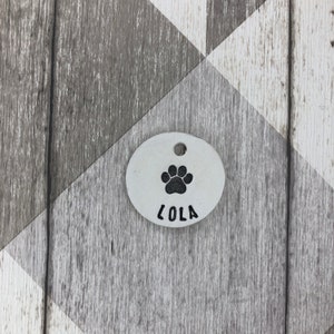 May include: White oval dog tag with a black paw print and the name "Lola" engraved on it.