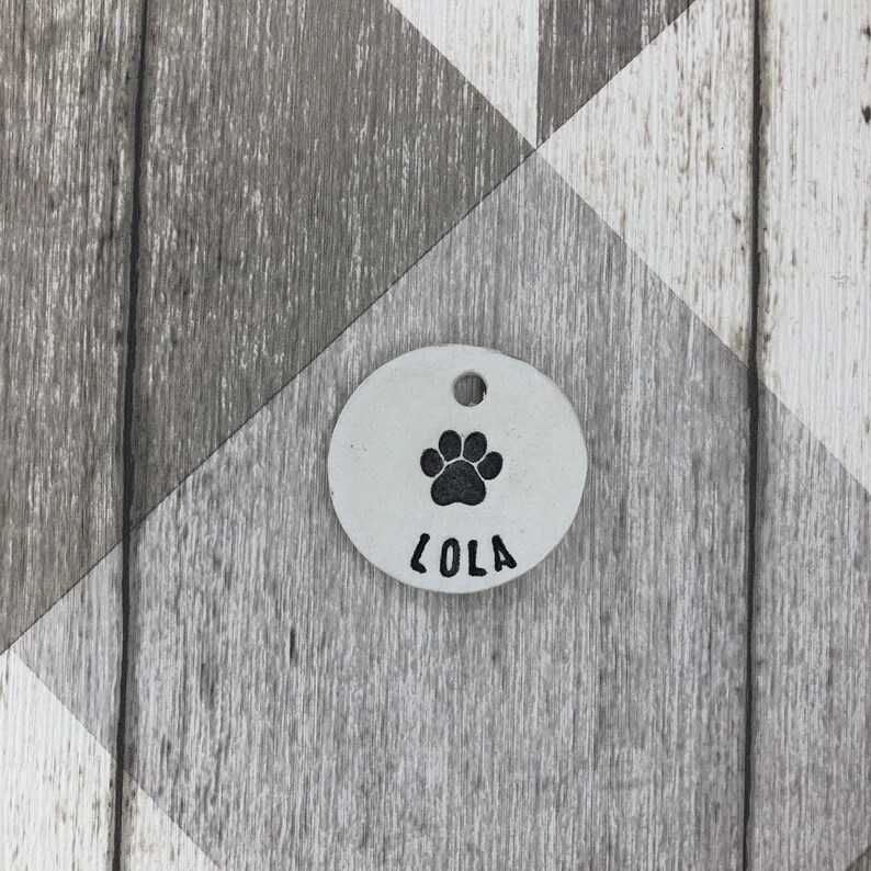 May include: White oval dog tag with a black paw print and the name "Lola" engraved on it.