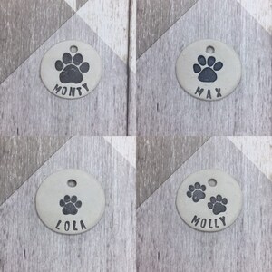 May include: Four round, light grey ceramic dog tags with black paw prints and names. The tags read "Monty", "Max", "Lola", and "Molly".