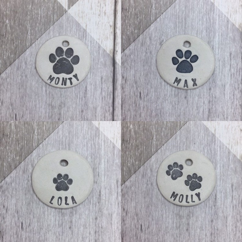 May include: Four round, light grey ceramic dog tags with black paw prints and names. The tags read "Monty", "Max", "Lola", and "Molly".
