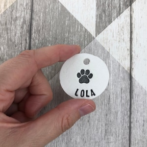 May include: White ceramic dog tag with a black paw print and the name "Lola" engraved on it.