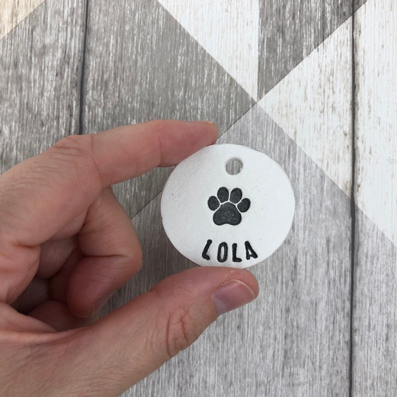 May include: White ceramic dog tag with a black paw print and the name "Lola" engraved on it.