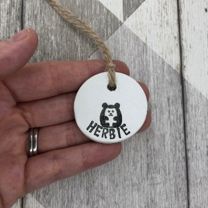 May include: A circular white ceramic ornament featuring a black hedgehog design and the name "HERBIE" beneath it. It hangs from a natural twine string. The background is a light grey and white wooden pattern.