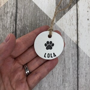 May include: White ceramic dog tag with a black paw print and the name "Lola" engraved on it. The tag is attached to a brown twine string.