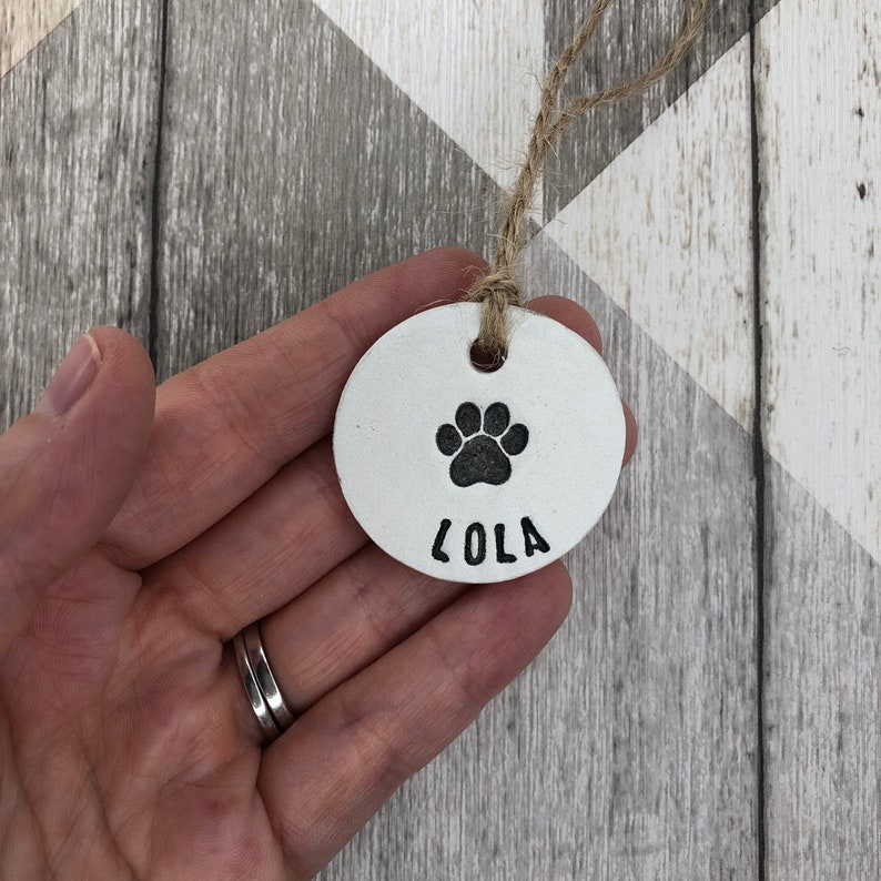 May include: White ceramic dog tag with a black paw print and the name "Lola" engraved on it. The tag is attached to a brown twine string.