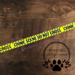 May include: Yellow crime scene tape with black text reading "CRIME SCENE DO NOT CROSS" diagonally across a wooden surface. The tape has a repeating pattern of black diagonal lines. A logo with a paw print and flowers is in the bottom right corner.