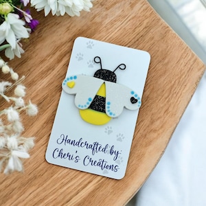 May include: A white and yellow bee with black stripes and blue accents. The bee is made of felt and has glitter on its wings. The bee is on a white card with the text "Handcrafted by: Cheri's Creations".
