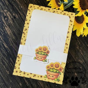 May include: A handmade greeting card with a yellow sunflower pattern and a white insert. The card features two stickers of smiling sunflowers in pots. The card is handcrafted by Cheri's Creations.