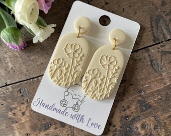 May include: A pair of cream-colored dangle earrings featuring an embossed floral design on oval-shaped pendants. The earrings are attached to a white display card with the text "Handmade with Love" and a small graphic of a cat holding a heart.