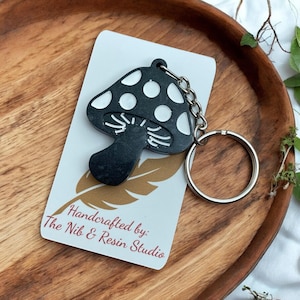May include: Black mushroom-shaped keychain with white polka dots and a silver keyring. The keychain is attached to a white card with the text "Handcrafted by: The Nib & Resin Studio" and a gold feather design. The keychain is on a wooden tray.