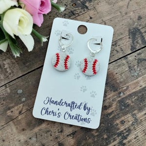 May include: A pair of silver-toned hoop earrings featuring circular charms designed to look like baseballs with white glittery surfaces and red stitching. The earrings are attached to a white display card printed with the text Handcrafted by: Cheri's Creations.
