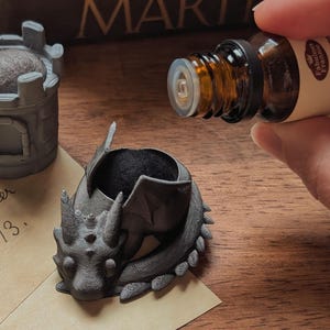 May include: A black dragon-shaped bottle stopper with a small, gray castle-shaped bottle stopper in the background. A bottle of essential oil is being held by a hand.