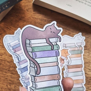 May include: A sticker of a brown cat sleeping on top of a stack of colorful books. The sticker is on a wooden surface.