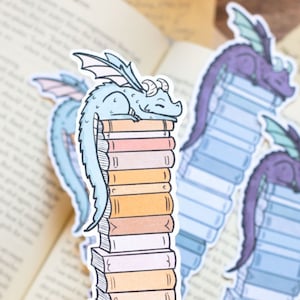 May include: Two colorful dragon bookmarks with a stack of books. One dragon is blue and the other is purple. Both dragons are sleeping on top of the books.