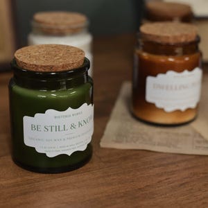 May include: A selection of glass candle jars with cork lids. The jars contain candles in shades of green and brown. Each jar has a white label with text. The front jar features the text "BE STILL & KNOW". The candles sit on a wooden surface.