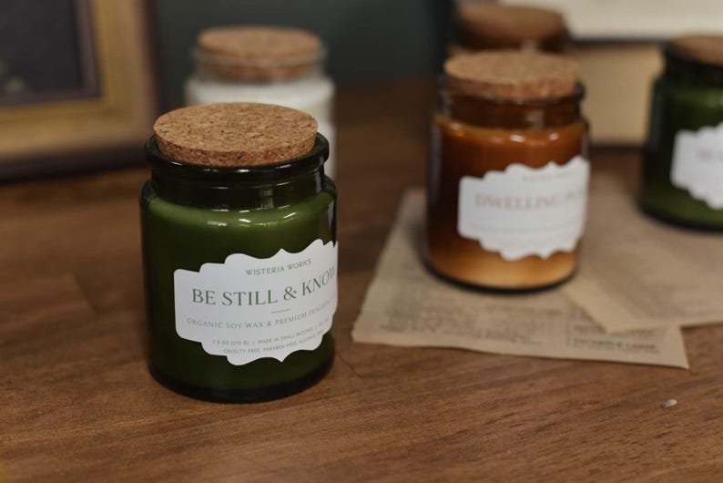 May include: A selection of glass candle jars with cork lids. The jars contain candles in shades of green and brown. Each jar has a white label with text. The front jar features the text "BE STILL & KNOW". The candles sit on a wooden surface.