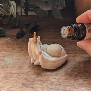 May include: White ceramic unicorn diffuser with a gold horn, holding a white diffuser ball. A hand is holding a brown glass bottle of essential oil over the diffuser ball.