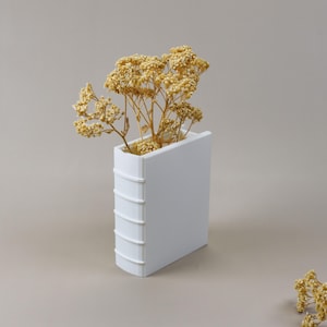 May include: A white, book-shaped vase holding dried yellow flowers. The vase has a rectangular shape with a spine detail, and the flowers add a natural element to the minimalist design. The vase is a decorative home accent.