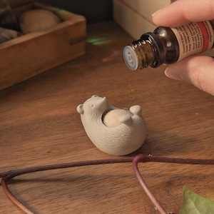 May include: A ceramic diffuser shaped like a hedgehog with a small white ball inside. A hand is holding a brown glass bottle with a dropper and a label that reads "Essential Oil".