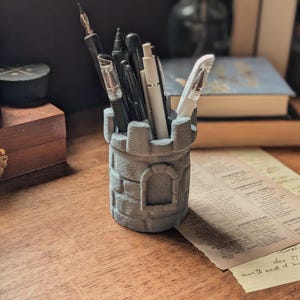 May include: A gray stone castle-shaped pencil holder with a variety of black and white pens and pencils inside.