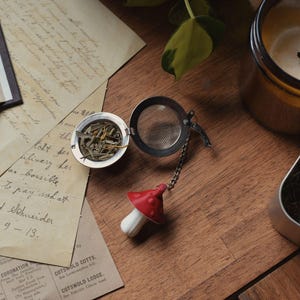 May include: A tea infuser with a red mushroom-shaped handle, open to reveal loose tea leaves. The infuser sits on a wooden surface with a candle, loose tea, and vintage papers.