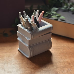 May include: A gray, book-shaped pencil holder with a stack of three books. The holder is filled with a variety of pens and pencils, including pink, black, and gray.
