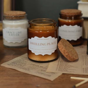 May include: A selection of glass jar candles with cork lids, in shades of green, brown, and white, are displayed. Each candle has a label with text. Matches and a piece of paper are in the foreground. The candles are on a wooden surface.