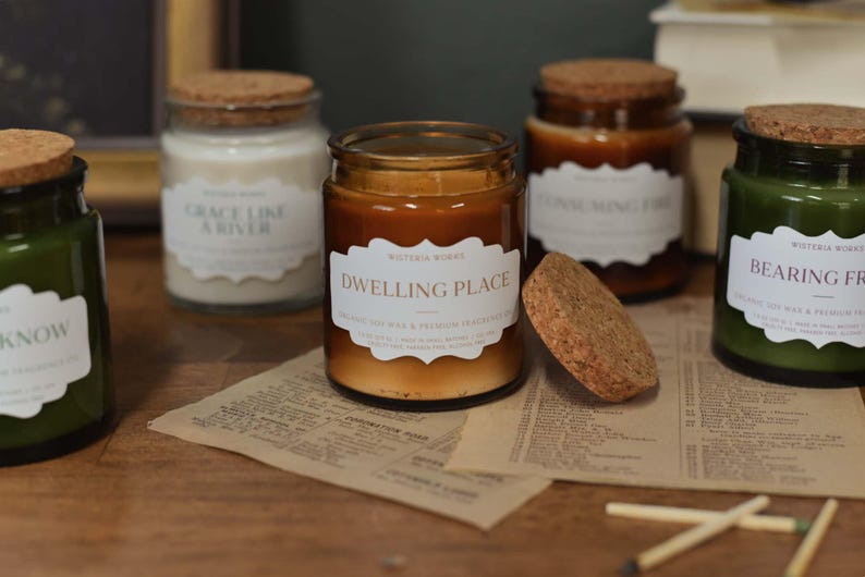 May include: A selection of glass jar candles with cork lids, in shades of green, brown, and white, are displayed. Each candle has a label with text. Matches and a piece of paper are in the foreground. The candles are on a wooden surface.