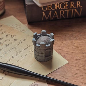 May include: A gray, castle-shaped pin cushion with a brown cushion top sits on a piece of paper with handwritten text. A black pen and a book by George R.R. Martin are also visible on the wooden surface.