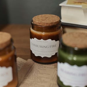 May include: A collection of glass candle jars with cork lids, in shades of brown and green, are displayed on a wooden surface. One jar features a label reading "CONSUMING FIRE". A book and newspaper are visible in the background.