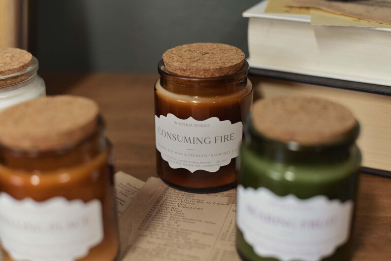 May include: A collection of glass candle jars with cork lids, in shades of brown and green, are displayed on a wooden surface. One jar features a label reading "CONSUMING FIRE". A book and newspaper are visible in the background.