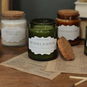 May include: A collection of glass jar candles with cork lids, in shades of green and amber, are displayed on a wooden surface. The labels on the candles feature phrases like "Be Still & Know" and "Bearing Fruit".