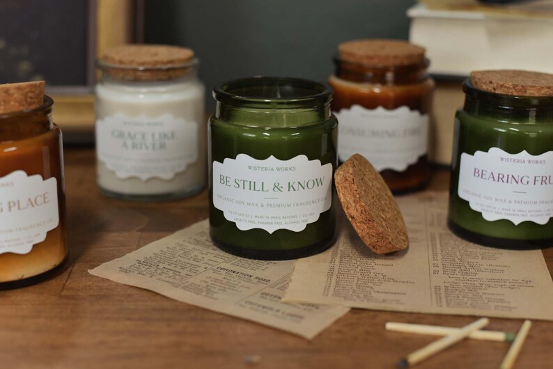 May include: A collection of glass jar candles with cork lids, in shades of green and amber, are displayed on a wooden surface. The labels on the candles feature phrases like "Be Still & Know" and "Bearing Fruit".
