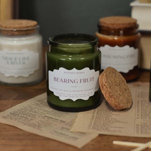 May include: A collection of glass jar candles with cork lids, in shades of green and brown, are displayed on a wooden surface. Each candle has a label; one reads "BEARING FRUIT". The scene includes a piece of paper with text and matches.