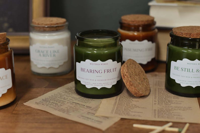 May include: A collection of glass jar candles with cork lids, in shades of green and brown, are displayed on a wooden surface. Each candle has a label; one reads "BEARING FRUIT". The scene includes a piece of paper with text and matches.