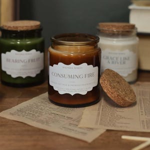 May include: A selection of scented candles in glass jars with cork lids. The labels read "Bearing Fruit", "Consuming Fire", "Grace Like a River", and "Be Still & Know". The candles are in shades of green, brown, and white, on a wooden surface.
