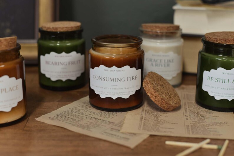 May include: A selection of scented candles in glass jars with cork lids. The labels read "Bearing Fruit", "Consuming Fire", "Grace Like a River", and "Be Still & Know". The candles are in shades of green, brown, and white, on a wooden surface.