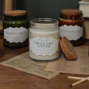 May include: A collection of scented candles in glass jars with cork lids. The labels on the jars read phrases such as "Grace Like a River" and "Bearing Fruit." The jars are in shades of green and brown, and are on a wooden surface.