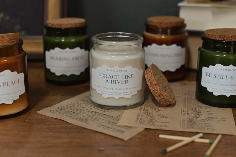 May include: A collection of scented candles in glass jars with cork lids. The labels on the jars read phrases such as "Grace Like a River" and "Bearing Fruit." The jars are in shades of green and brown, and are on a wooden surface.