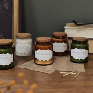 May include: Five glass jar candles with cork lids and labels. The candles are in shades of green, white, and brown. The labels read "Be Still & Know", "Grace Like a River", "Dwelling Place", "Final Mingling Fire", and "Bearing Fruit".